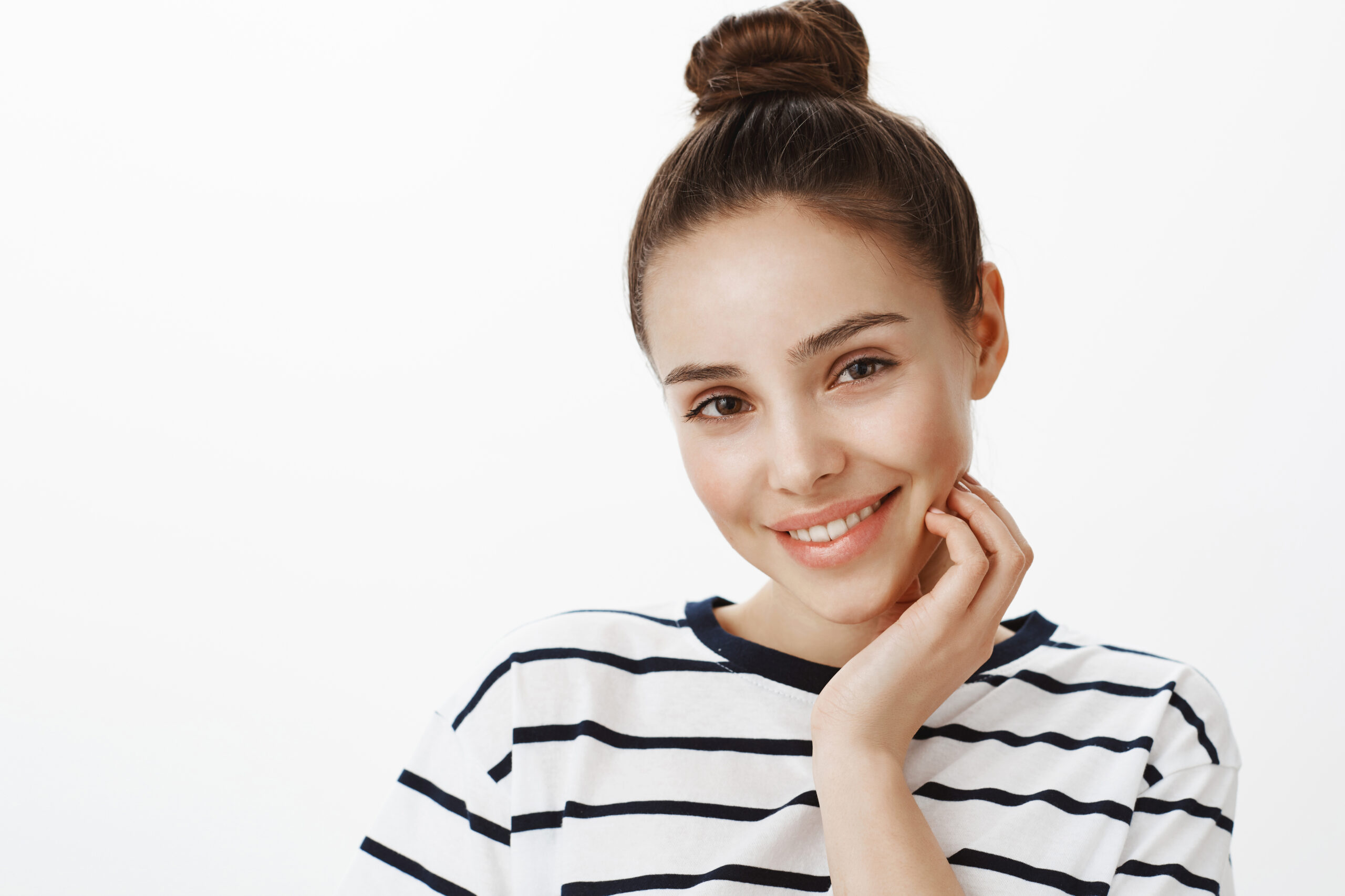 Studio shot of confident attractive european female with stylish haircut in striped outfit, touching face gently and smiling sensually at camera, flirting and being in romantic mood over white wall.