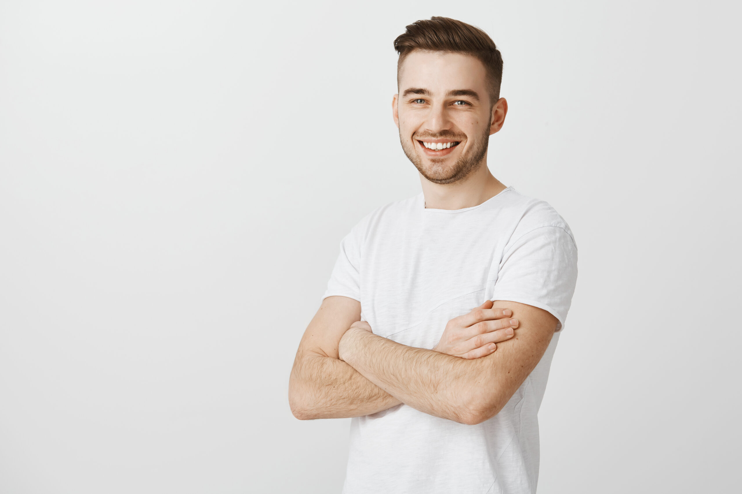 Portrait of pleased confident and accomplished european guy with stylish haircut standing unshaven over gray background crossing hands on chest smiling joyfully and satisfied at camera.