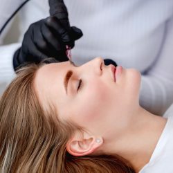 Face of girl in profile during microblading permanent makeup process. Hands of tattoo master holding professional ink machine with medicine needle