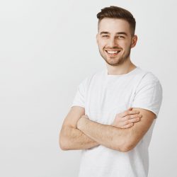 Portrait of pleased confident and accomplished european guy with stylish haircut standing unshaven over gray background crossing hands on chest smiling joyfully and satisfied at camera.