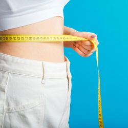 Slim woman measures her waist waistline with measuring tape. Healthy body shaping weight loss concept. Slim waist small belly in big white denim pants isolated over blue color background. Copy space.